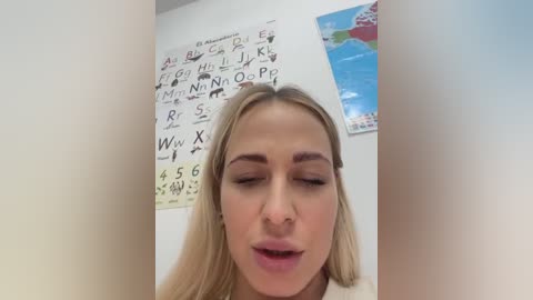 Media: Video of a young blonde woman with closed eyes, possibly in a classroom setting, with whiteboard posters and colorful maps in the background.