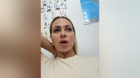 Media: Video of a blonde woman with fair skin, wearing a beige top, posing indoors. Background includes a wall map and educational posters.