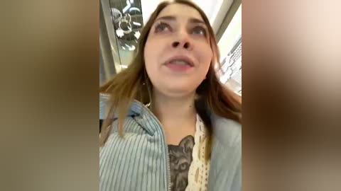 Media: A video of a young woman with light skin, long brown hair, and a striped shirt, looking up with a slightly open mouth, taken indoors with blurred background.