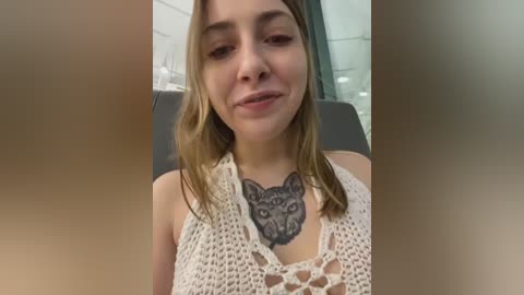 Media: Video of a young woman with fair skin, medium-length blonde hair, wearing a white crocheted top. She has a tattoo of a cat's face on her chest. Background shows a blurred modern interior with white tiles and a window.