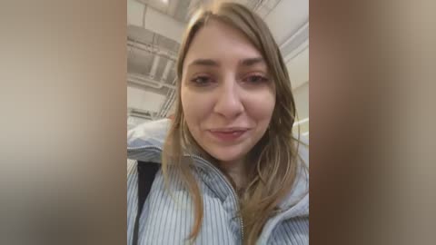 Media: Video of a young woman with light skin and straight, shoulder-length brown hair, wearing a light blue, striped shirt. Background shows an industrial room with exposed pipes and white walls.