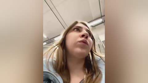 Media: A candid video of a young woman with fair skin and light brown hair, wearing a light blue jacket, captured from a low angle in a dimly lit room with white ceiling tiles.