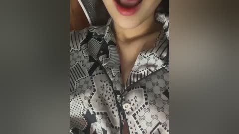 Media: Video of a woman's face and chest, wearing a silver satin blouse with a geometric black and white pattern. She has light skin and full lips painted with a red lipstick.