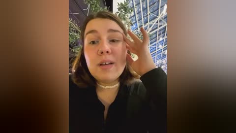 Media: Video of a young woman with fair skin, shoulder-length brown hair, and a black choker necklace, wearing a black top, smiling, with a plant in the background.