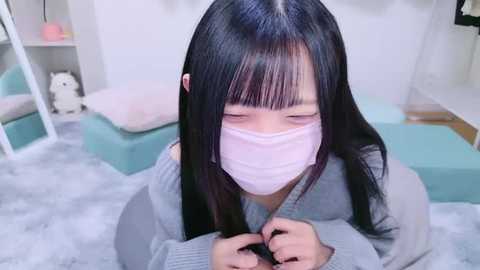 Media: A video of an East Asian woman with long black hair and a pale complexion, wearing a light pink surgical mask, standing in a pastel-colored, softly lit room with white walls, a fluffy carpet, and a mirror.