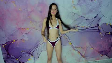 Video of a slender, light-skinned woman with long black hair, wearing a red bikini with white trim, standing against a pastel, abstract, marble-patterned wall.