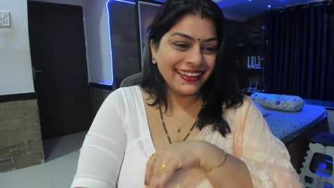 Media: Video of a smiling South Asian woman in a white saree and gold necklace, holding a baby in a dimly lit bedroom with a bed and dark curtains in the background.