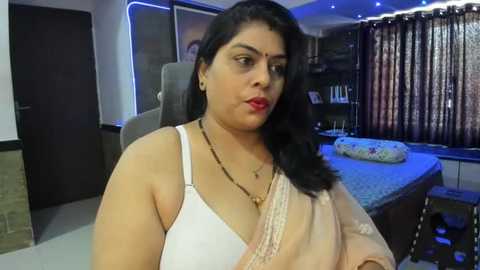 Media: Video of a South Asian woman with medium complexion, dark hair, and fair skin, wearing a white bra and beige sari, seated in a modern living room with a white door, dark curtains, and a flower-patterned pillow.