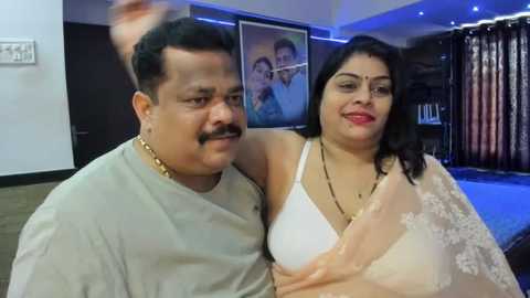 Media: Video of a middle-aged South Asian couple, man in gray shirt and gold chain, woman in white blouse, sitting closely in a modern living room with dark walls, blue lighting, and framed family photos.