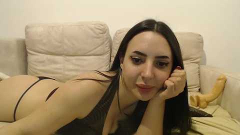 Media: Video of a young woman with long dark hair lying on a beige couch, wearing a black thong and black top, with a dildo and a stuffed banana beside her.