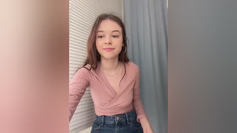 Video of a young Caucasian woman with light skin and brown hair, wearing a pink ribbed V-neck top and high-waisted blue jeans, standing in a dressing room with white blinds and gray curtains.