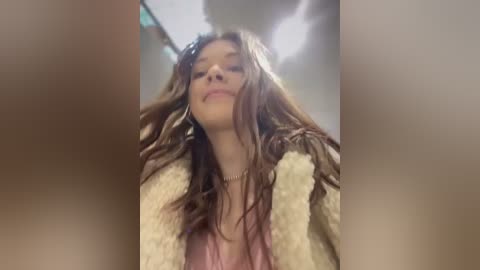 Video of a young woman with long, wet, brown hair, wearing a beige, fluffy bathrobe, standing in a dimly lit bathroom with a cloudy mirror.