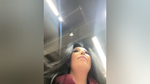 Media: Video of a woman with long black hair, fair skin, and maroon lipstick, looking upwards in a dimly lit, modern interior with bright fluorescent lights.