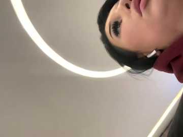 Media: A video of a woman with long, straight black hair, fair skin, and dark eyeliner, wearing a maroon sweater, looking upward. The background features a circular, bright white light fixture against a plain, light-colored ceiling.