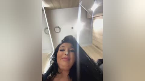 Media: A video of a woman with long black hair, fair skin, and large breasts, smiling, standing in a modern, bright room with wooden floors and minimalist decor.