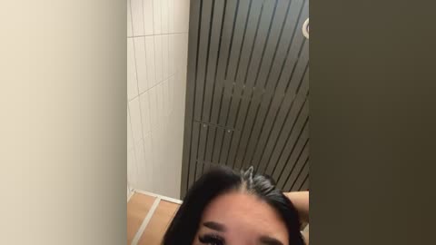 Media: A video of a woman with long black hair, taken from an overhead angle, showing her face and upper torso. She is standing in a tiled bathroom with beige walls and a metallic ceiling.