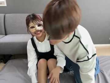 Media: Video of two children, a girl with a white dress and black pinafore, and a boy in a sweater, wearing masks, sitting on a gray couch in a modern living room.