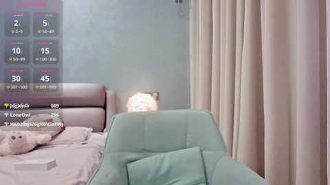 Media: Video of a modern bedroom with a light blue upholstered armchair, a beige headboard, and a soft, pink lampshade. The background features pale grey walls and light pink curtains. A digital clock overlay displays time and weather.