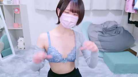 Media: Video of an Asian woman with short black hair, wearing a light blue lace bra, grey cardigan, and face mask, dancing energetically in a pink, white, and blue-themed room with plush toys and a grey couch.