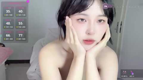 Media: A young East Asian woman with short black hair, fair skin, and a slim build, sits topless, resting her head on her hands, in a softly lit room. A digital thermometer overlay shows her temperature at 77.0\u00b0F.