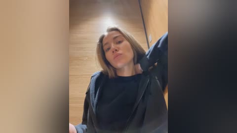 Media: Video of a young Caucasian woman with light skin and shoulder-length blonde hair, wearing a black leather jacket and black top, leaning against a wooden wall in a dimly lit hallway.