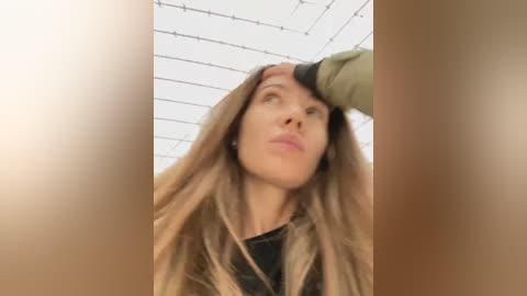 Video of a woman with long, straight brown hair, wearing a black top, looking up at a hand in an enclosed, white-tiled room with a grid ceiling.