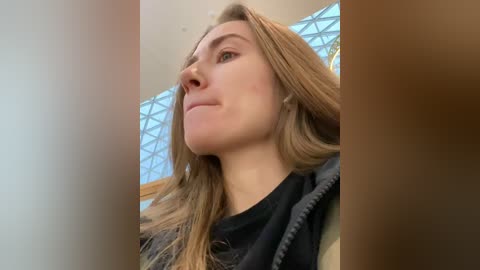 Video of a Caucasian woman with light skin and long blonde hair, wearing a black top, standing indoors near a glass atrium with geometric patterns.