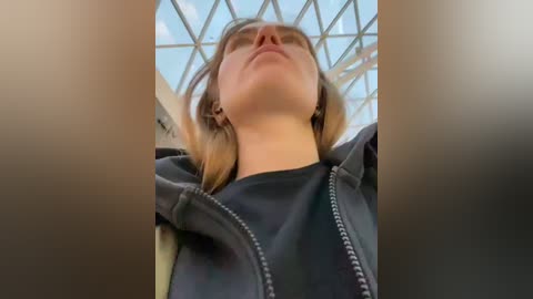 Video of a young woman with light skin and straight, shoulder-length blonde hair, wearing a black hoodie, captured from a low angle. The background features a glass and metal ceiling structure with a bright blue sky visible through the glass.