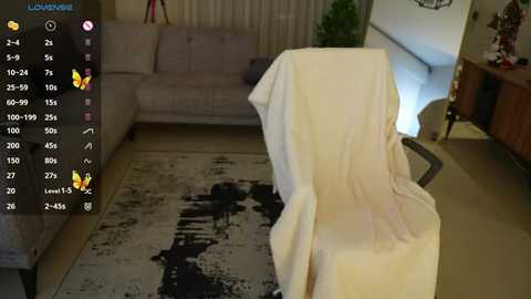 Media: Video of a dimly-lit living room with a beige sofa, a draped chair, and a textured black-and-white rug. The background includes a potted plant and a wooden cabinet.