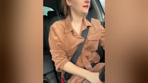 Media: A video shows a woman with light skin, brown hair, and red lipstick, wearing a beige, button-up shirt, seated in a car with a black seatbelt across her chest.