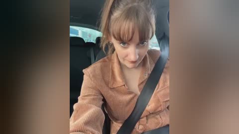 Media: Video of a young Caucasian woman with fair skin, light brown hair in a ponytail, wearing a ribbed brown sweater, buckled into a car seat, looking intently ahead.