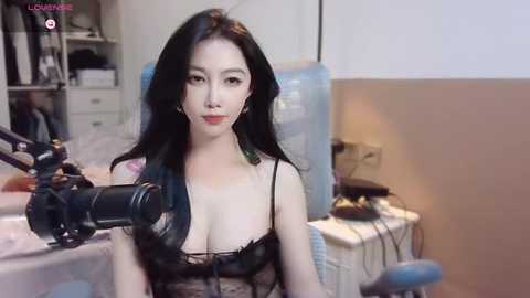 Media: Video of an East Asian woman with long black hair, wearing a revealing black lace bra, seated at a desk with a camera, in a modern, sparsely furnished room.