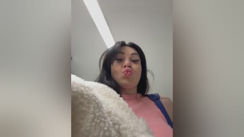 Media: Video of an Asian woman with long black hair, wearing a pink and blue sleeveless dress, blowing a kiss into the camera from a tight, white-walled corridor.