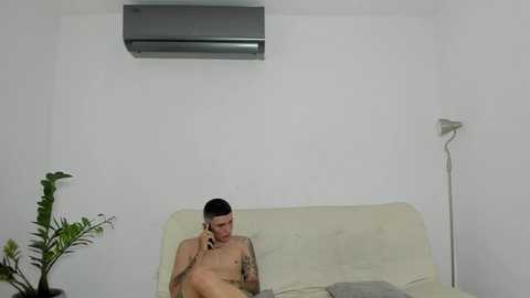 Media: Video of a naked man with short dark hair, medium build, and tattoos on his arms, sitting on a light-colored couch in a sparsely decorated room with a white wall, air conditioner, and lamp.