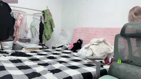 Media: Video of a messy bedroom with a bed covered in a black-and-white checkered blanket, clothes hanging on a rack, a pink cushion, and a green chair in the foreground.