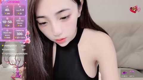 Media: A video of an East Asian woman with long dark hair, wearing a black halter top, sitting on a beige couch, with a smartphone displaying social media statistics in the background.