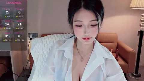 Media: Video of an Asian woman with black hair tied back, wearing a see-through white blouse revealing cleavage, sitting in a modern living room with a beige couch, lamp, and a TV screen displaying weather and temperature.