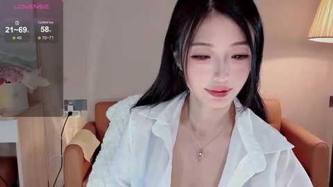 Video of an Asian woman with long black hair, wearing a white blouse, sitting indoors. She has fair skin and is smiling softly.