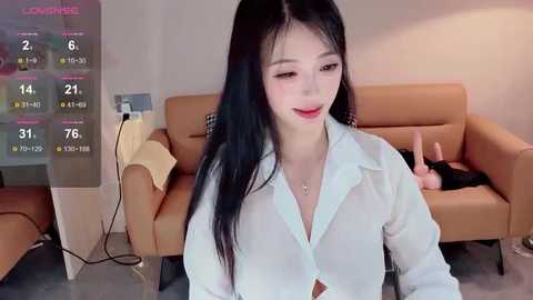 Media: A video shows an East Asian woman with long black hair, fair skin, and red lipstick, wearing a white blouse, seated in a modern living room with beige furniture and a digital clock.