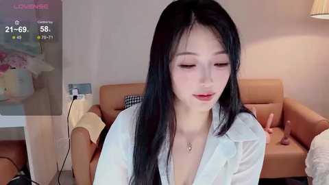 Video of an East Asian woman with long black hair, wearing a white blouse, sitting in a beige armchair in a modern living room.