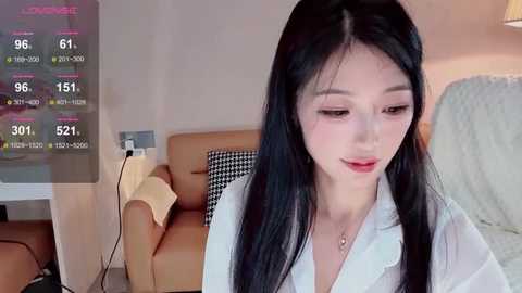 Media: Video of an East Asian woman with long black hair and fair skin, wearing a white blouse, sitting on a brown couch in a modern living room with a white rug and a digital thermometer display.