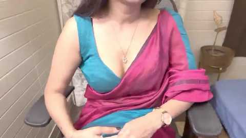 Media: Video of a light-skinned woman with shoulder-length dark hair, wearing a revealing turquoise and pink sari, seated in a medical examination room with white brick walls and a beige lamp.