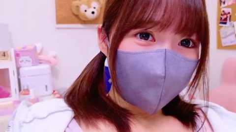 Media: Video of a young East Asian woman with straight brown hair in pigtails, wearing a gray face mask, pink shirt, and white jacket. Background features a pastel room with stuffed toys and a corkboard.