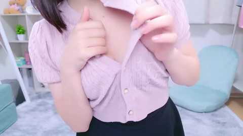 Media: Video of an East Asian woman with light skin, wearing a pink button-up shirt, black skirt, and beige panties, posing in a pastel-colored room with white shelves, plants, and a turquoise chair.