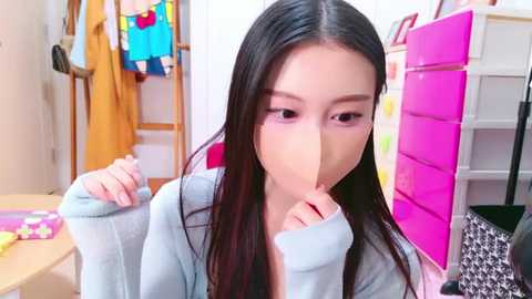 Media: Video of an Asian woman with long black hair, wearing a light blue hoodie, covering her mouth with her hand, in a colorful, organized room with a pink locker and clothing racks.