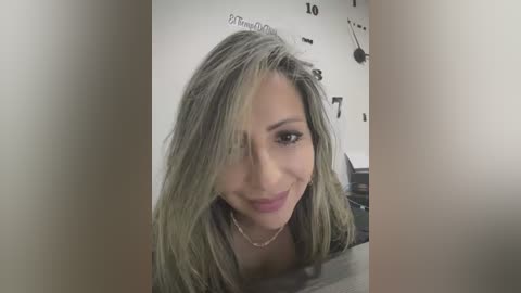 Media: Video of a smiling, light-skinned woman with long, wavy blonde hair, wearing a black top, in a room with a white wall clock and clock face.