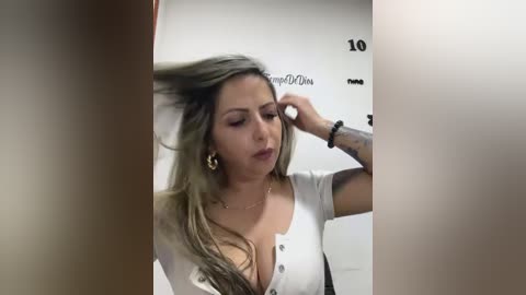 Media: A video shows a light-skinned woman with long, wavy, blonde hair, wearing a low-cut white top, adjusting her hair in front of a mirror.