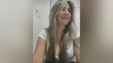 Media: Video of a young, Caucasian woman with long, wavy, light brown hair and a medium-sized bust, wearing a white t-shirt. She's smiling and has tattoos on her arm. The background is a plain, beige wall.