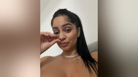 Media: Video of a smiling, topless woman with medium brown skin, long black hair tied back, and a pearl necklace. She playfully touches her nose with her finger. Background is a plain, off-white wall.