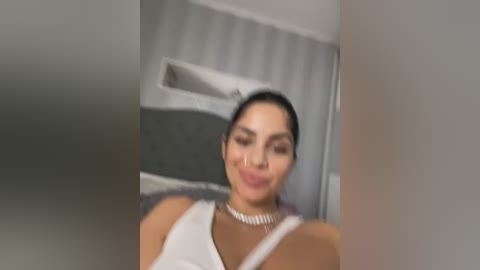 Media: A blurred video of a Latina woman with dark hair and medium skin tone, wearing a white dress and necklace, standing in a modern room with vertical blinds.